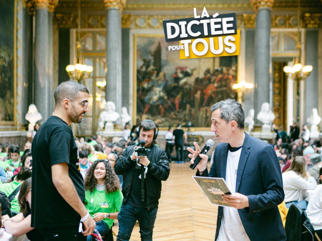 Two people stand in an ornate French palace hall during 'La Dictee Pour Tous' event with camera crew filming.