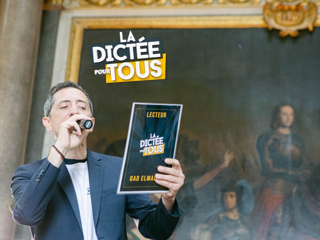 Person in a suit enthusiastically holds up a book titled 'La Dictee Pour Tous' while speaking into a microphone.