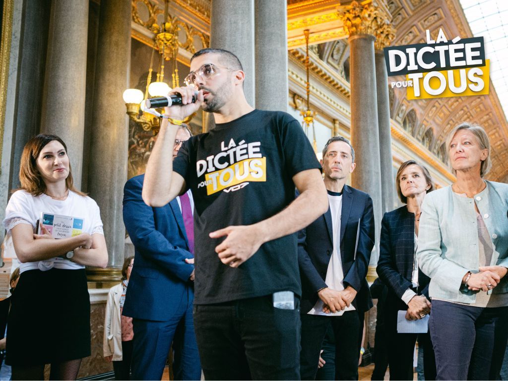 People gather to watch as someone speaks into a microphone while wearing a black Dictee t-shirt near columns.