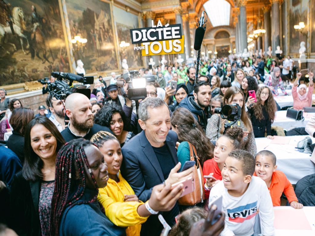 Diverse crowd gathers in ornate hall with large paintings during Dictee Tous event in Paris.