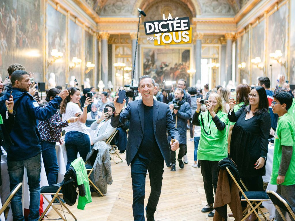 Crowd gathers in a grand palace hall for the Dictee Pour Tous spelling event with green-themed decorations.