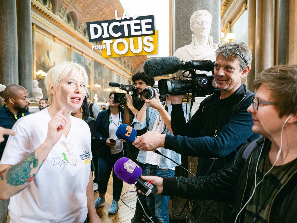 TV camera operators and reporters gather around an enthusiastically speaking person during a 'Dictee Tous' media event.
