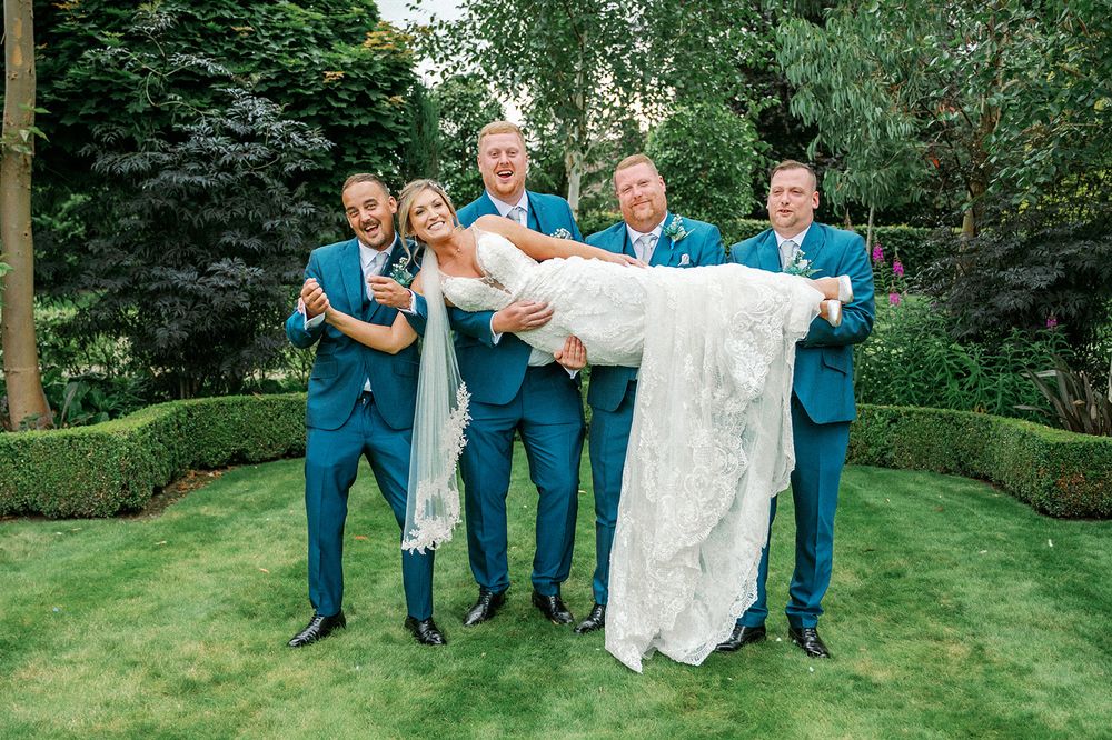 Wedding party in blue suits and white dress posing playfully on green lawn.