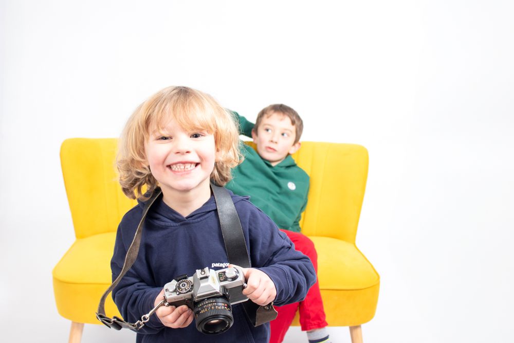 Child holding camera while sitting on yellow couch against white background.