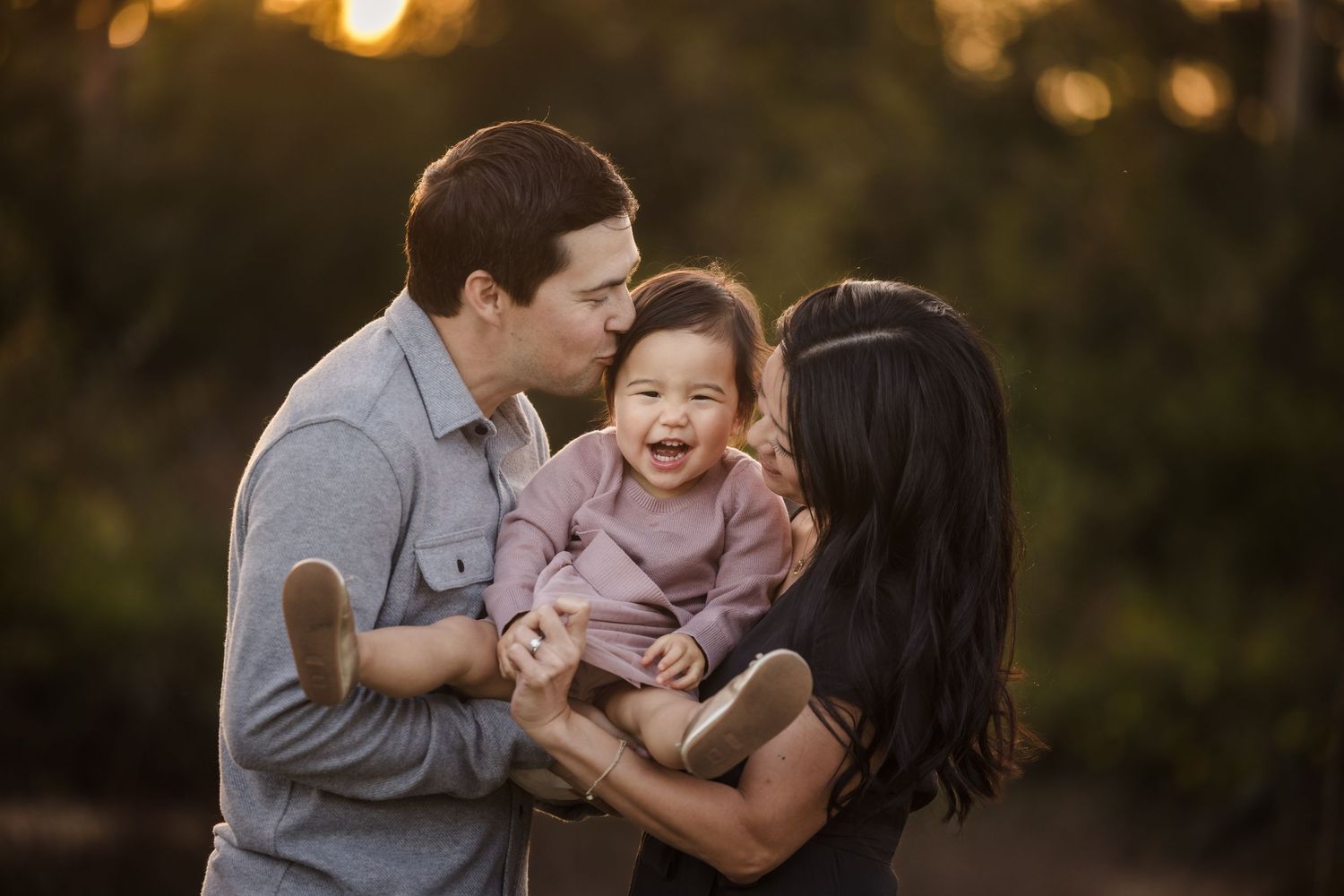 Parents hold and play with their laughing child during a warm sunset photoshoot outdoors.
