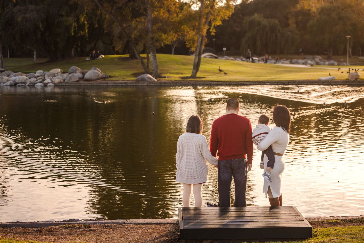 Three people stand together on a wooden dock overlooking a serene lake at sunset in a park setting.