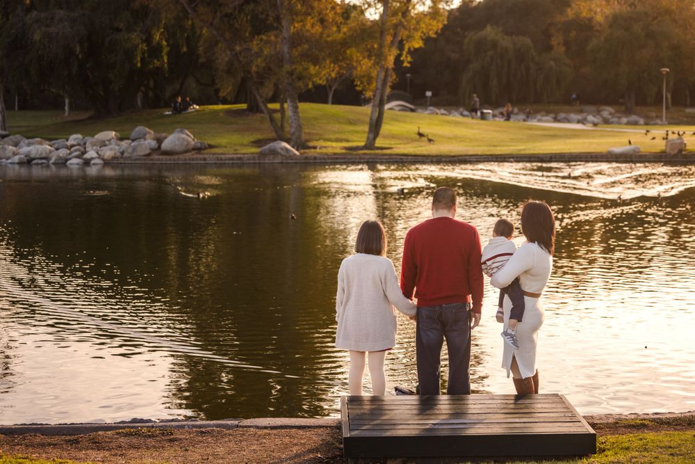 Three people stand together on a wooden dock overlooking a serene lake at sunset in a park setting.
