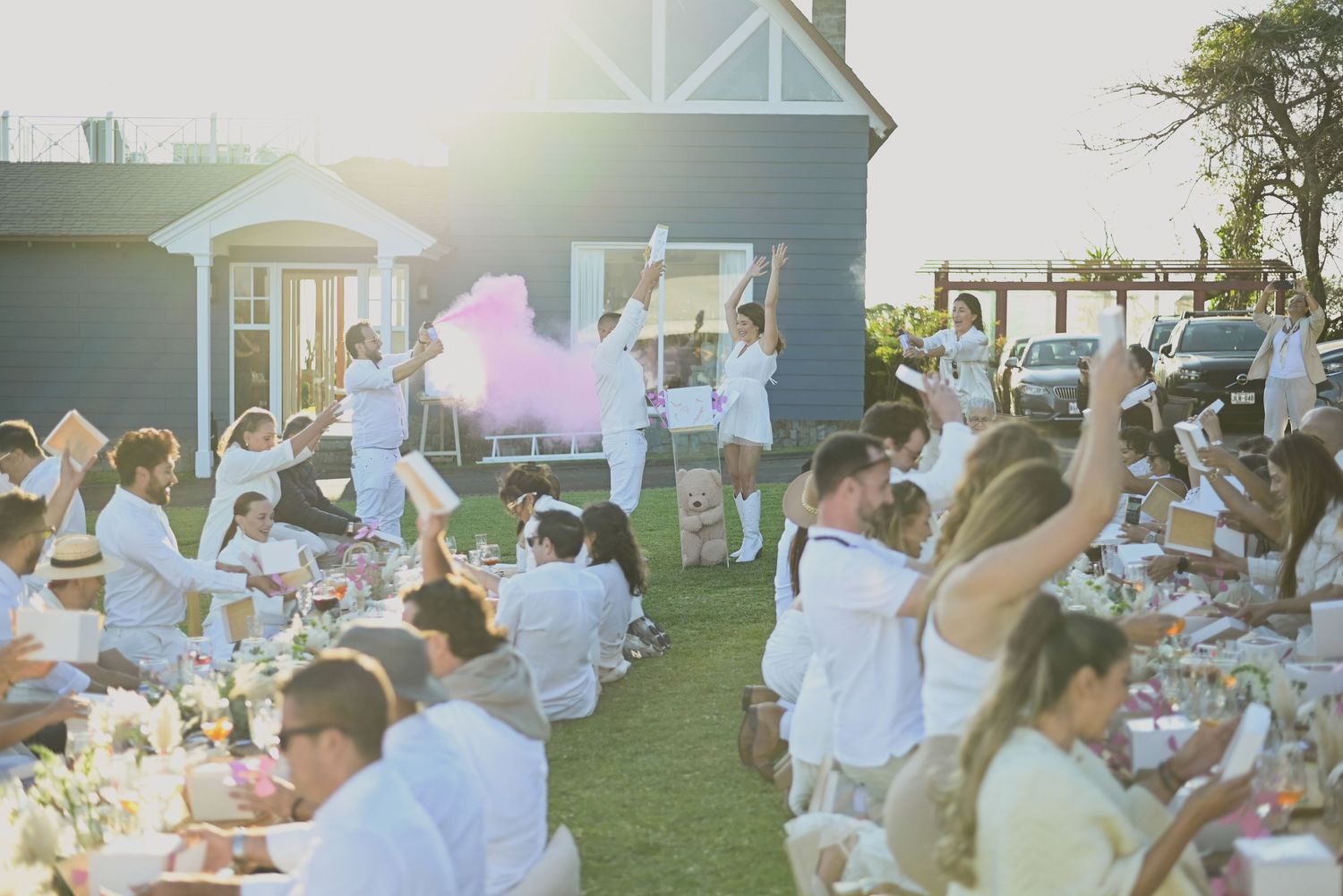 Outdoor evening dinner party with guests seated at long tables on a lawn as golden sunlight illuminates the scene.