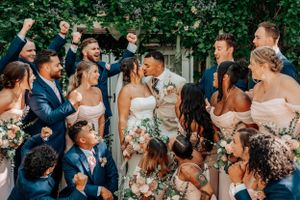 A joyous wedding party celebrates outdoors with the bride and groom in the center surrounded by bridesmaids and groomsmen.