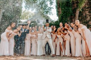 A wedding party in blush pink and beige formal attire celebrates with champagne spray outdoors under trees.