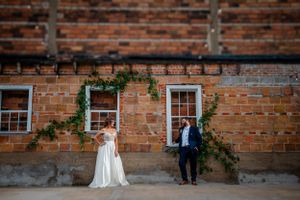 Wedding couple stands against rustic brick wall with climbing vines and white windows.
