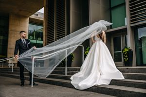 A beautiful wedding veil flows dramatically in the wind as a couple stands on modern building steps in elegant attire.