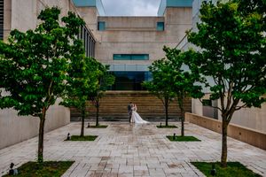 Modern building walkway lined with symmetrical green trees and stone pavers leads to entrance on overcast day.