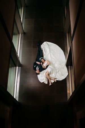 Overhead view of two people in formal attire laying on dark tiled floor with flowing white dress spread out.