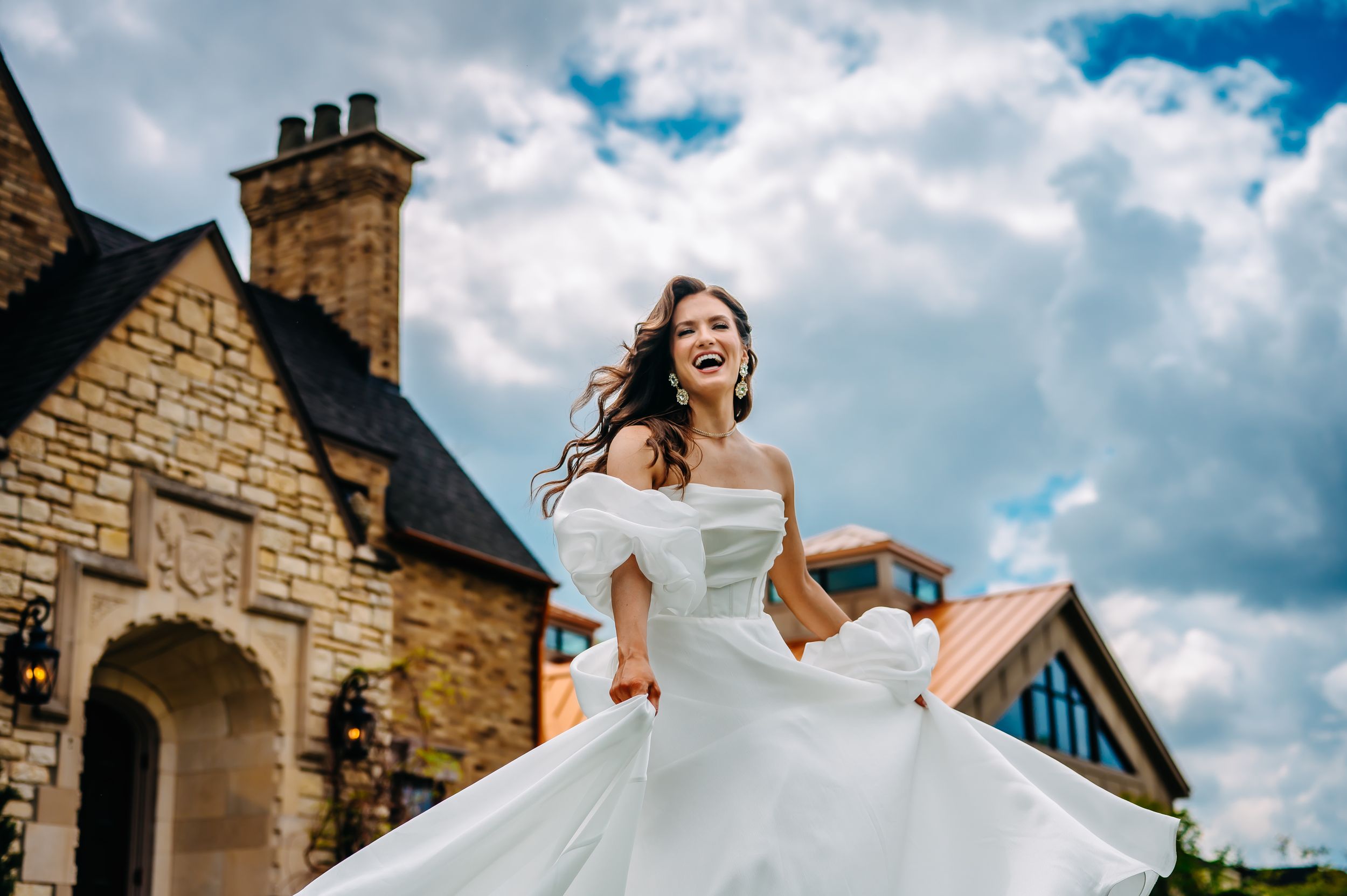 A joyful bride in a flowing white dress twirls in front of a stone house under blue skies with fluffy clouds.