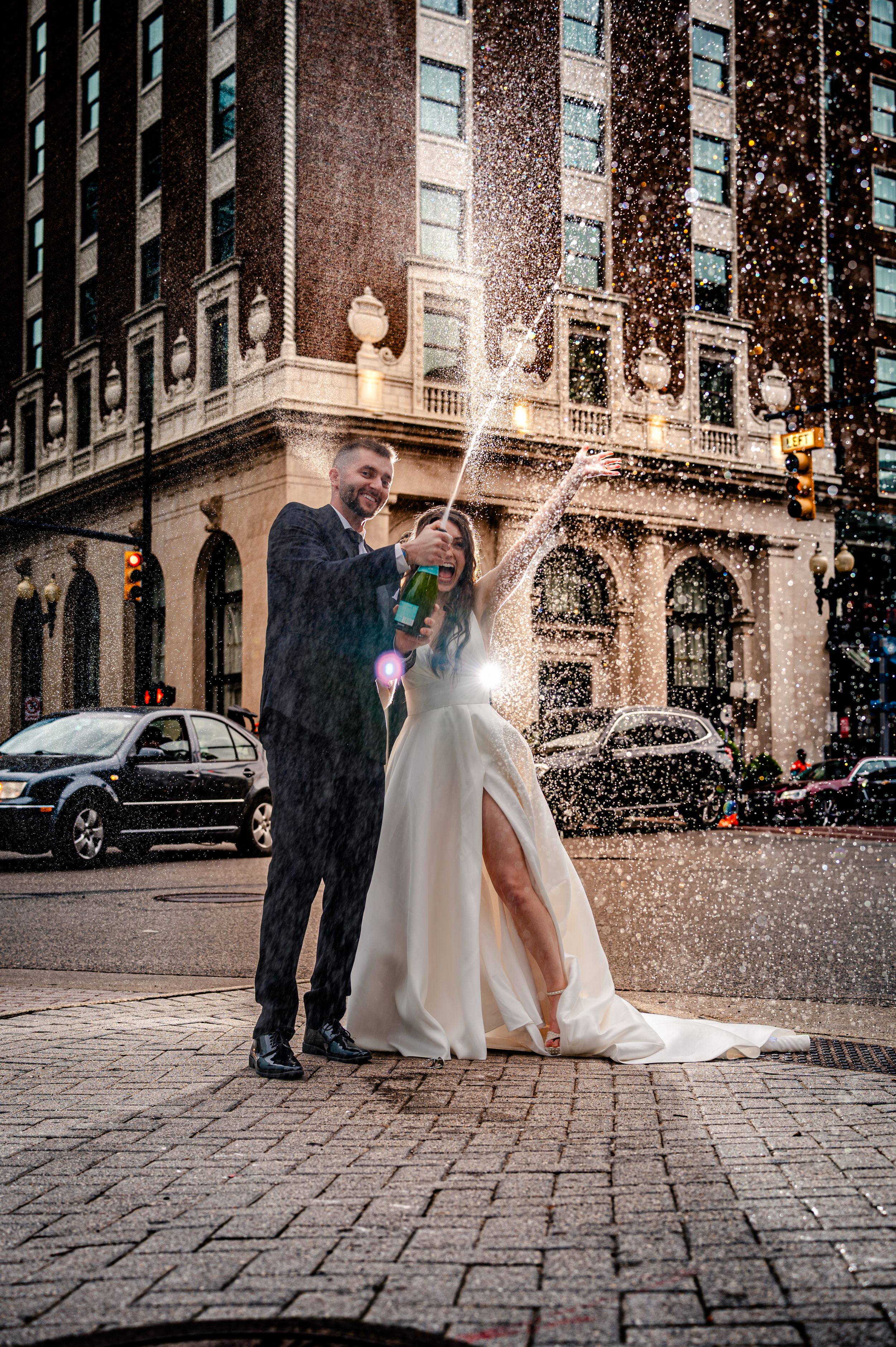 A couple pops champagne on a cobblestone street corner outside a historic building during a wedding celebration.