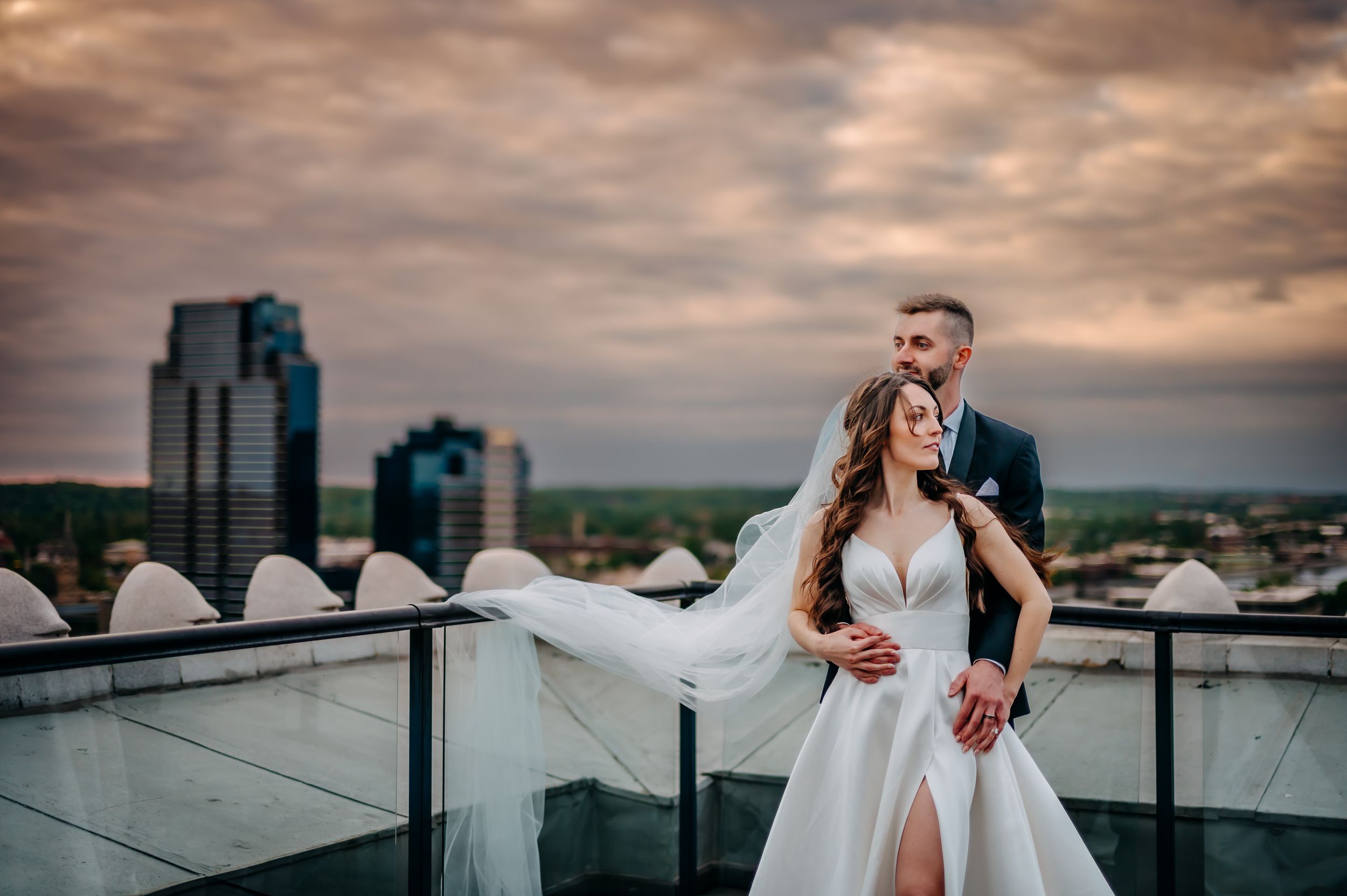 A romantic wedding couple embraces on a rooftop terrace with dramatic cloudy skies and city skyline at sunset.