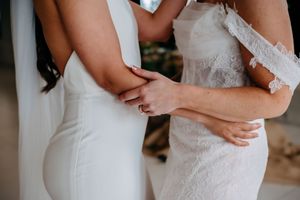Two people in elegant white wedding dresses hold hands in an intimate moment during their special day.
