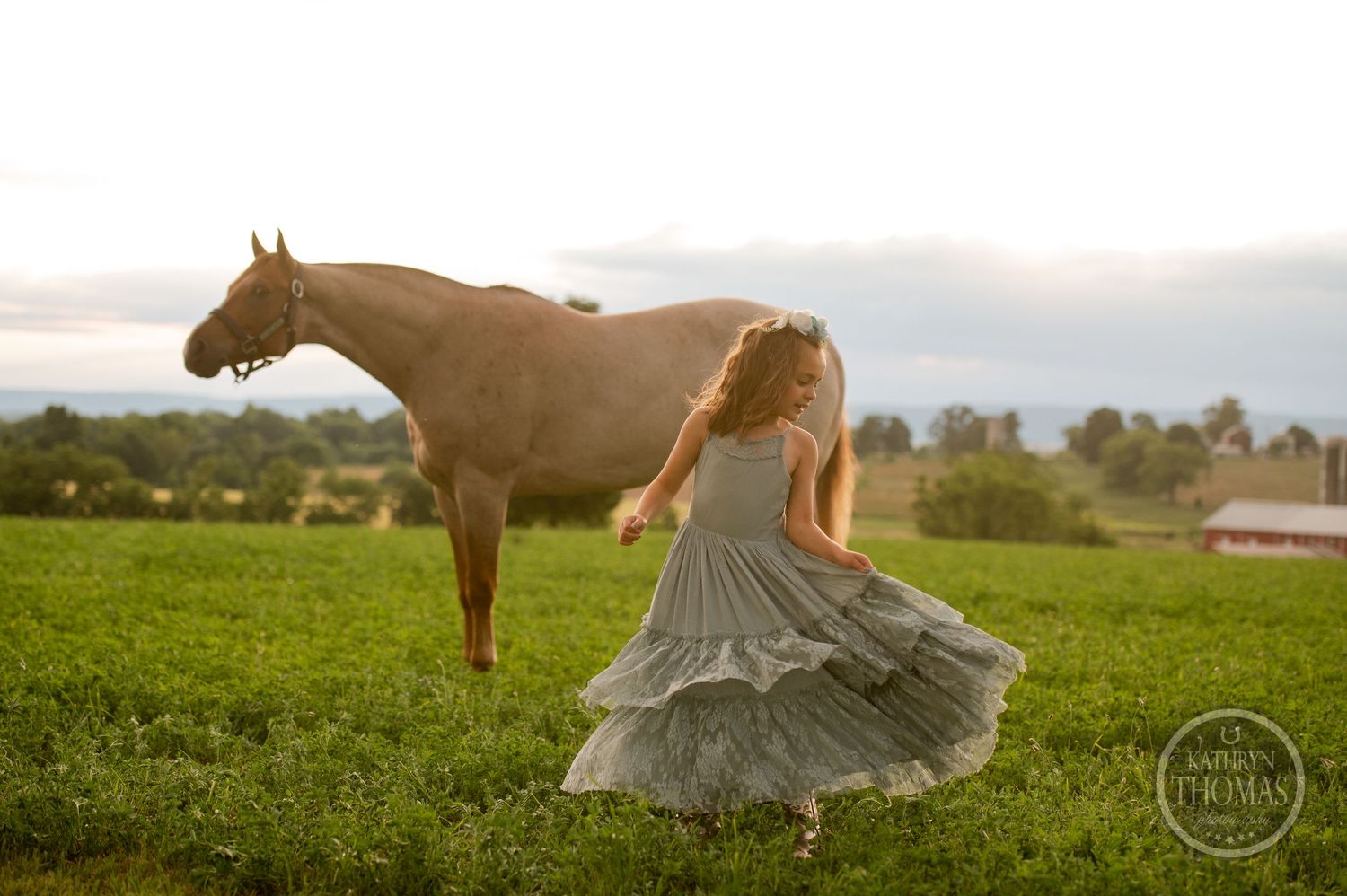 Figure in flowing grey dress dances in field beside grazing horse during sunset.