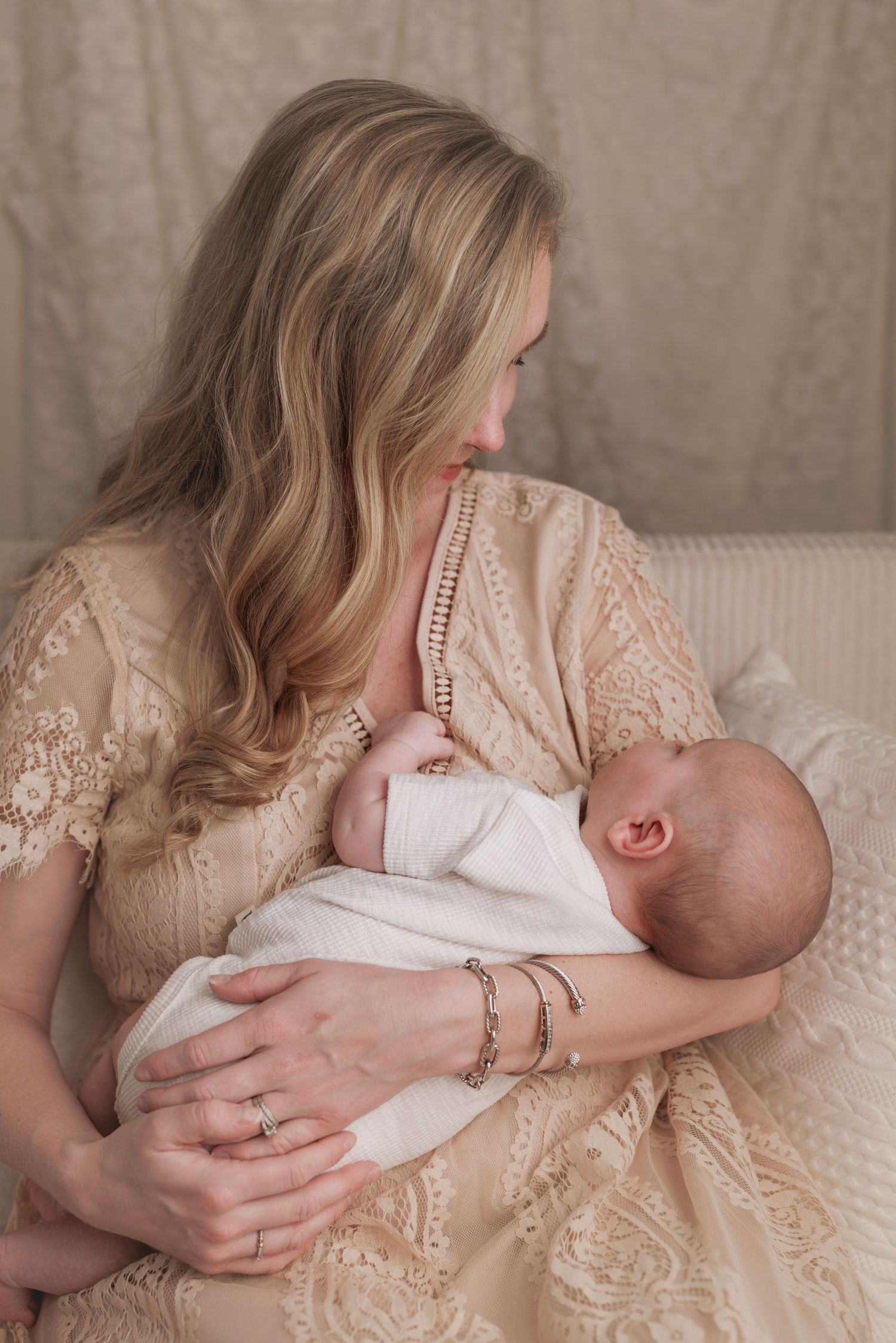 Mother in delicate lace dress breastfeeds newborn baby while sitting on beige couch.