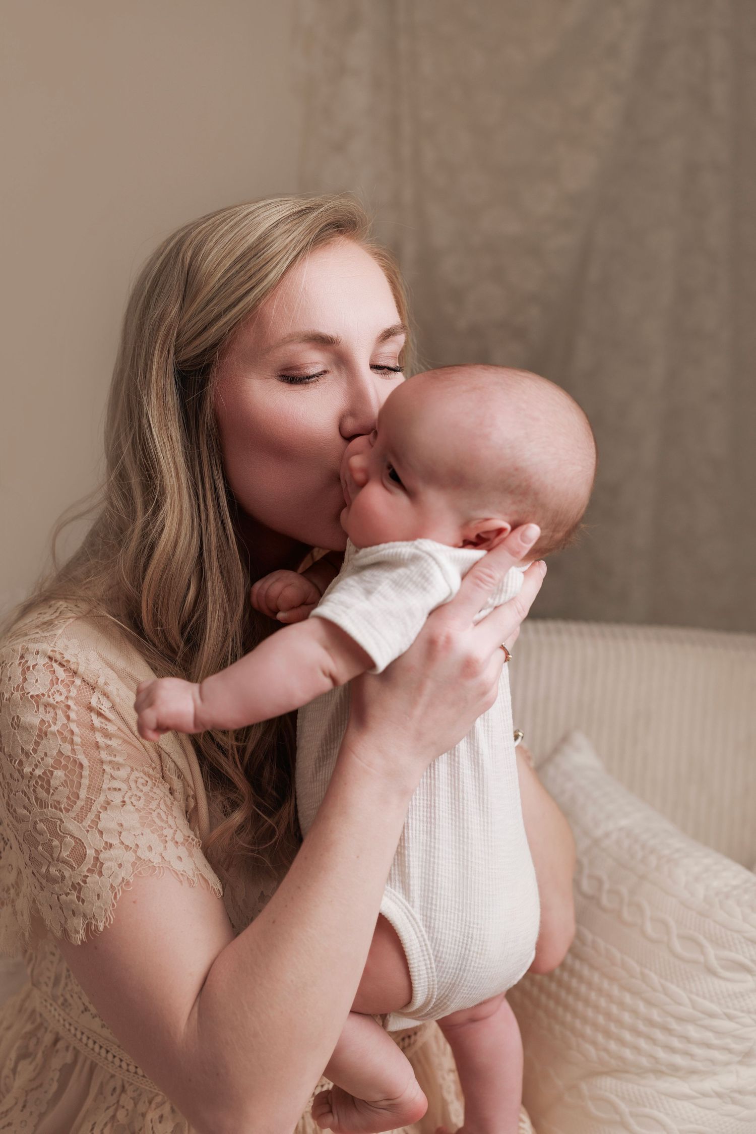A tender moment between a mother wearing a lace dress and her baby in white, sitting on a beige couch.