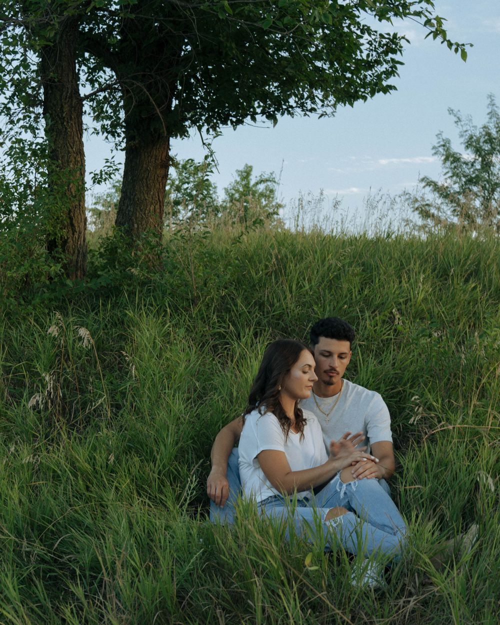 Couple sitting together in tall grass under tree during sunset.