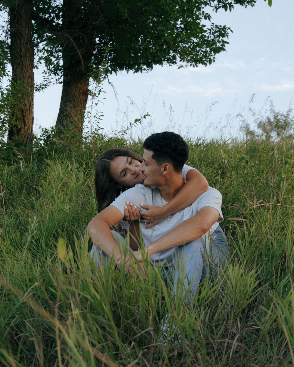 Couple embracing and laughing together in a grass field near a tree.