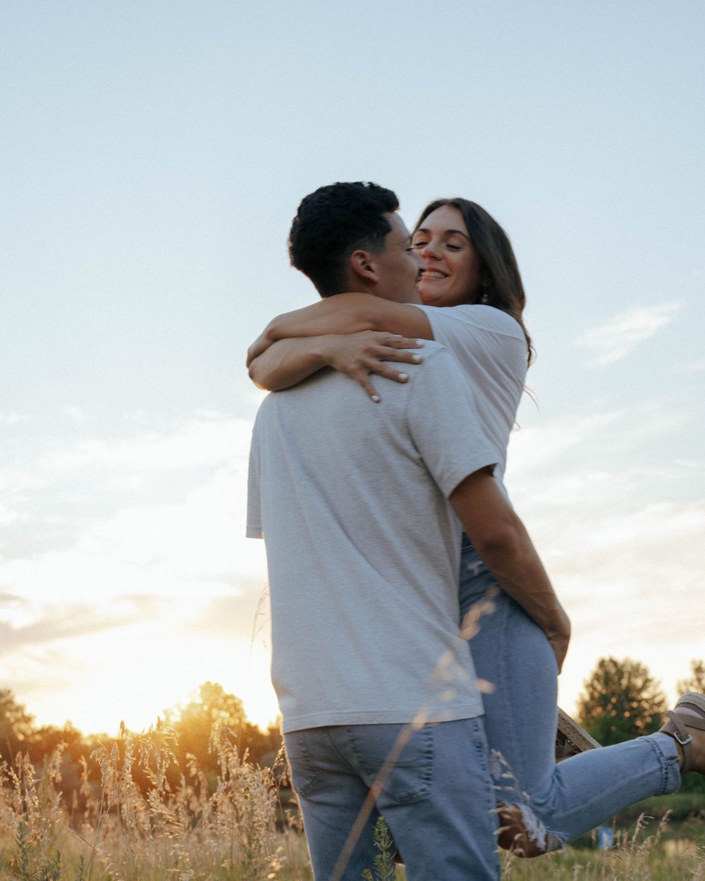 Romantic embrace outdoors during golden hour in a field with sunlit trees in background.