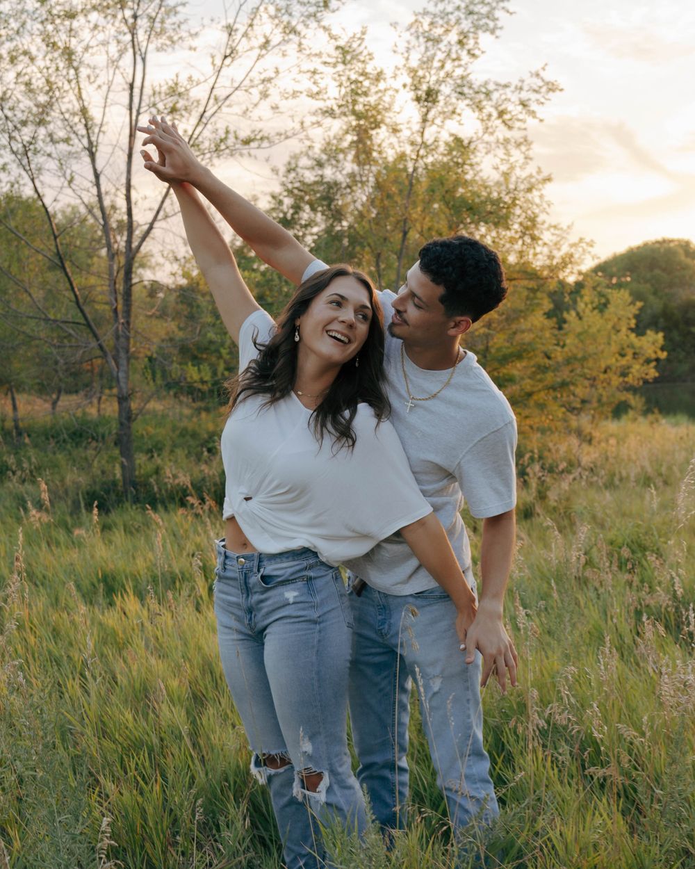 A couple dancing and laughing together in a sunlit field with tall grass and trees in the background.