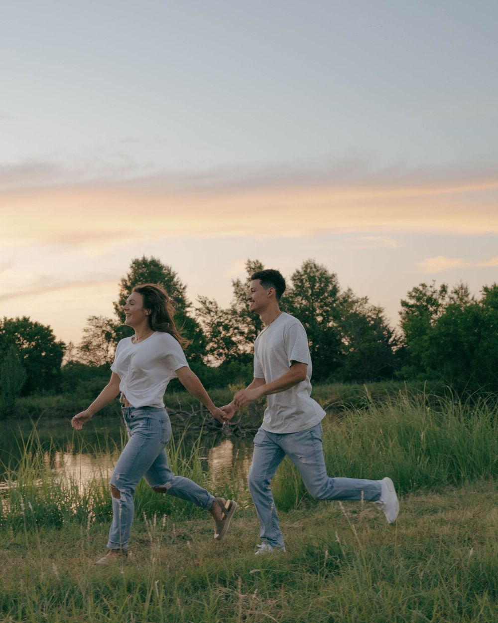 A couple running playfully through a grassy field at sunset.
