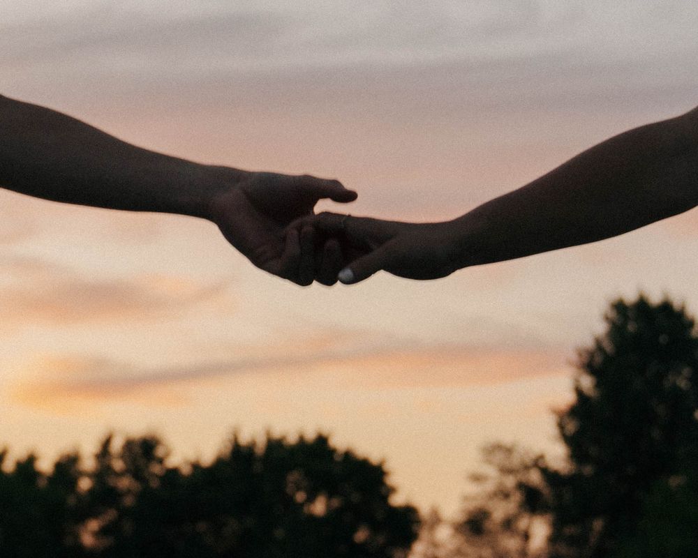 Silhouette of two hands reaching toward each other against an orange sunset sky.