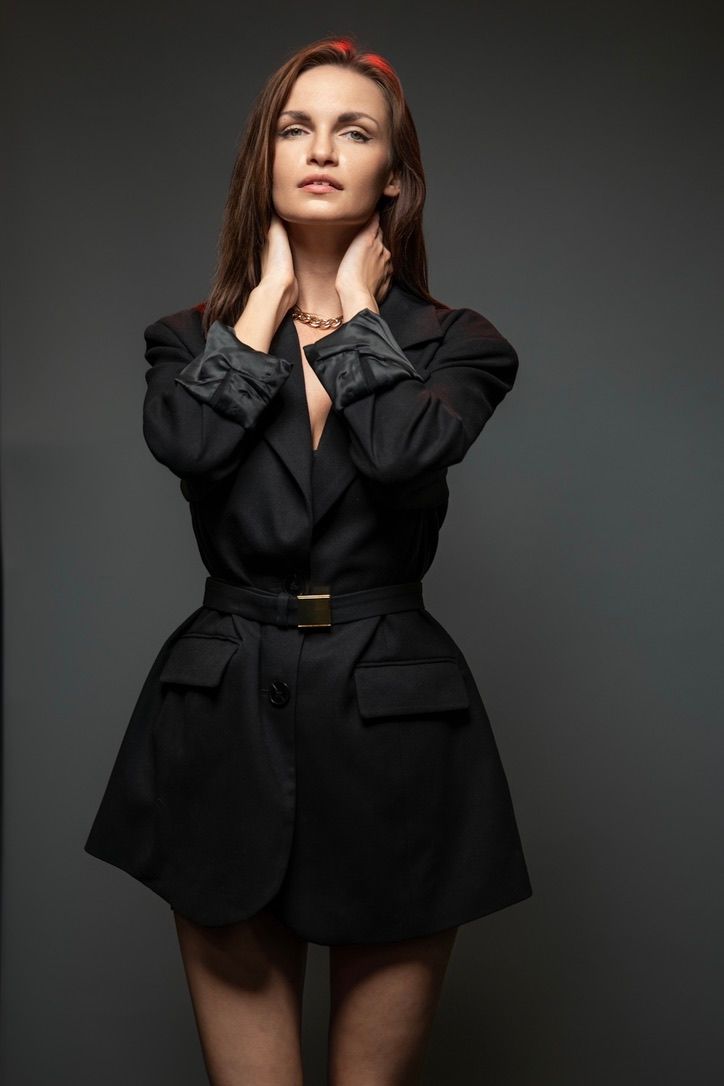 Fashion portrait of woman in black belted dress jacket against dark background.