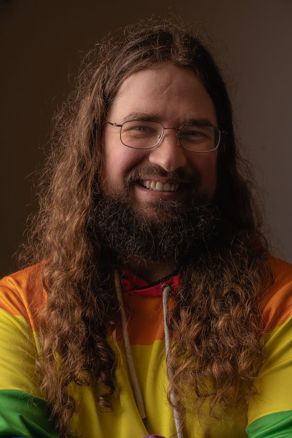 Person with long beard and hair smiles wearing colorful rainbow striped shirt.
