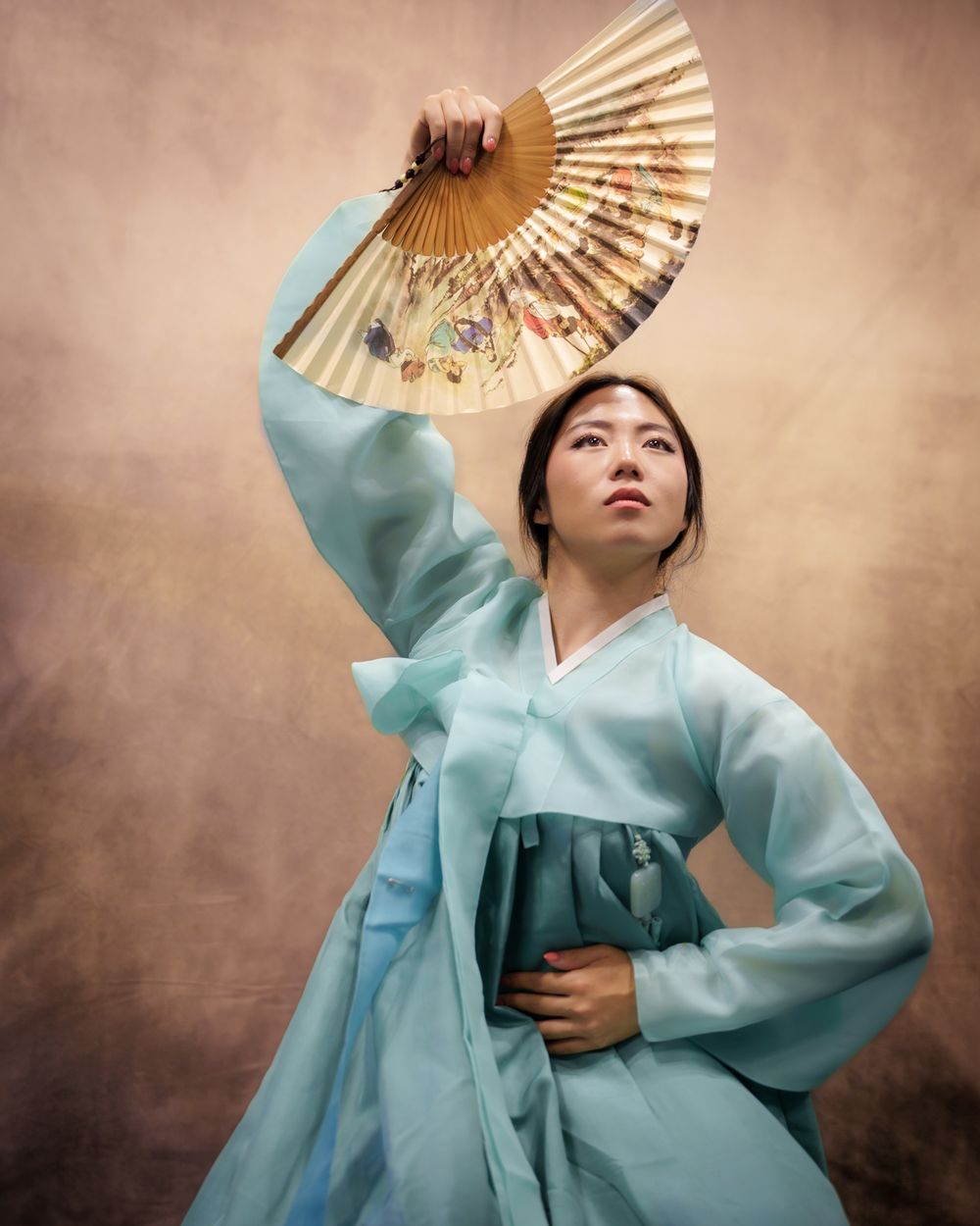 Woman in light blue hanbok holding decorative fan in artistic portrait.