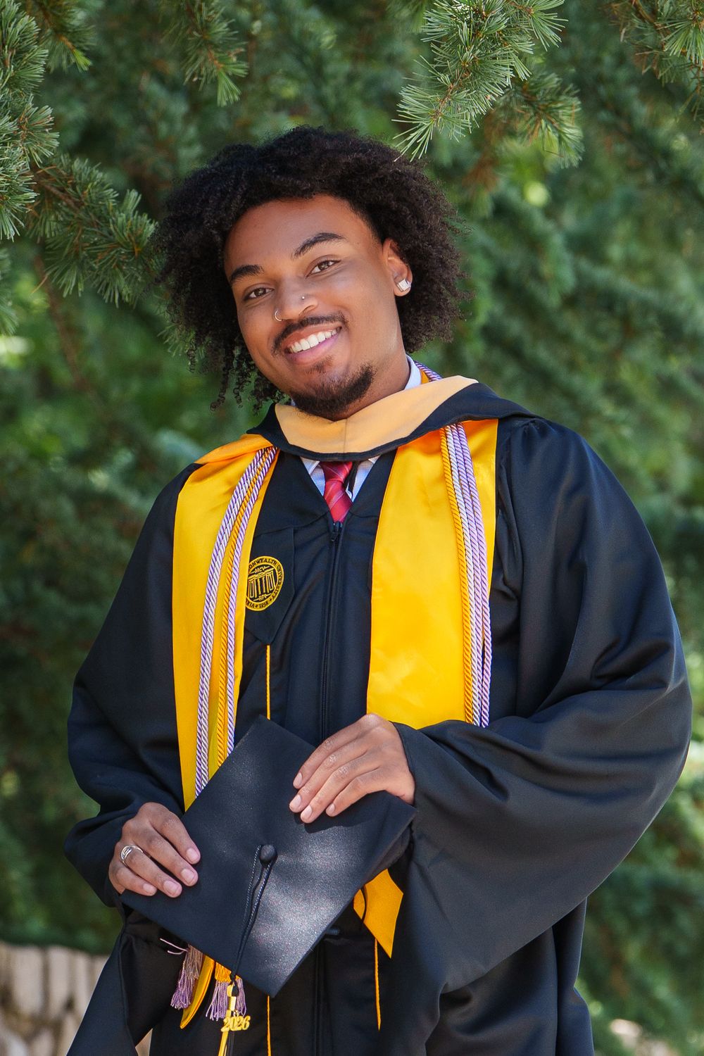 Smiling graduate in black and gold regalia holds cap outdoors near pine trees.