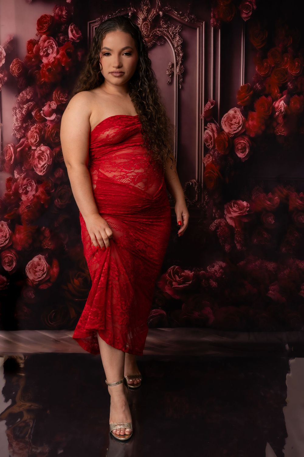 Woman in elegant red lace dress poses against dark floral backdrop in romantic portrait session.