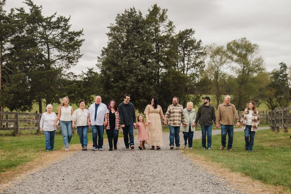 Large family group holding hands walking down a gravel path surrounded by green trees and farmland.