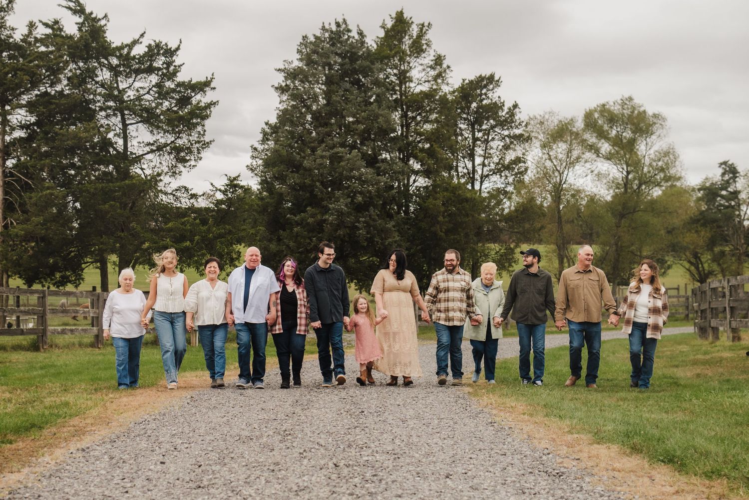 Large family group holding hands walking down a gravel path surrounded by green trees and farmland.