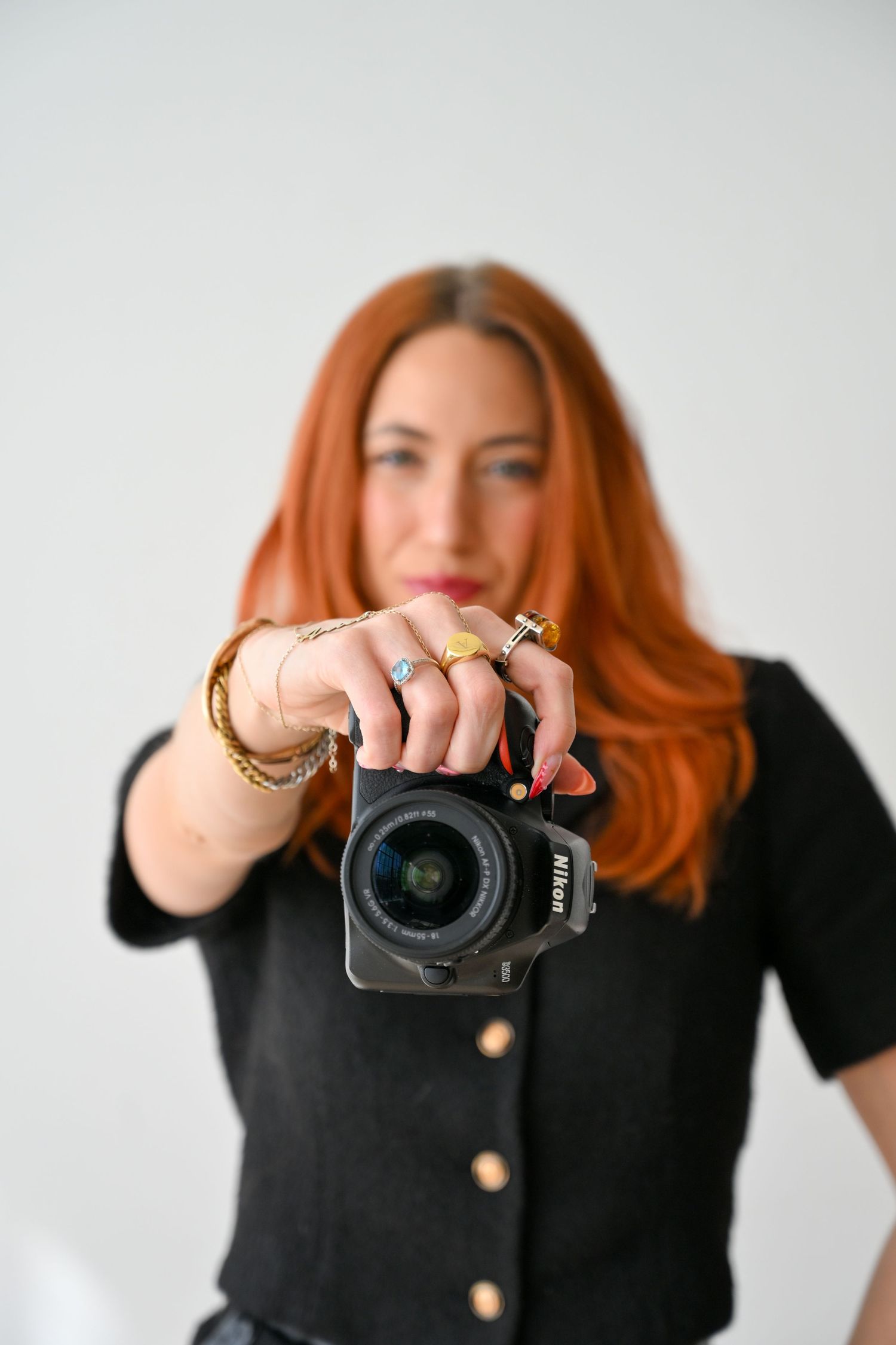 A woman with vibrant red hair wearing a black tank top and denim smiles warmly against a gray background.