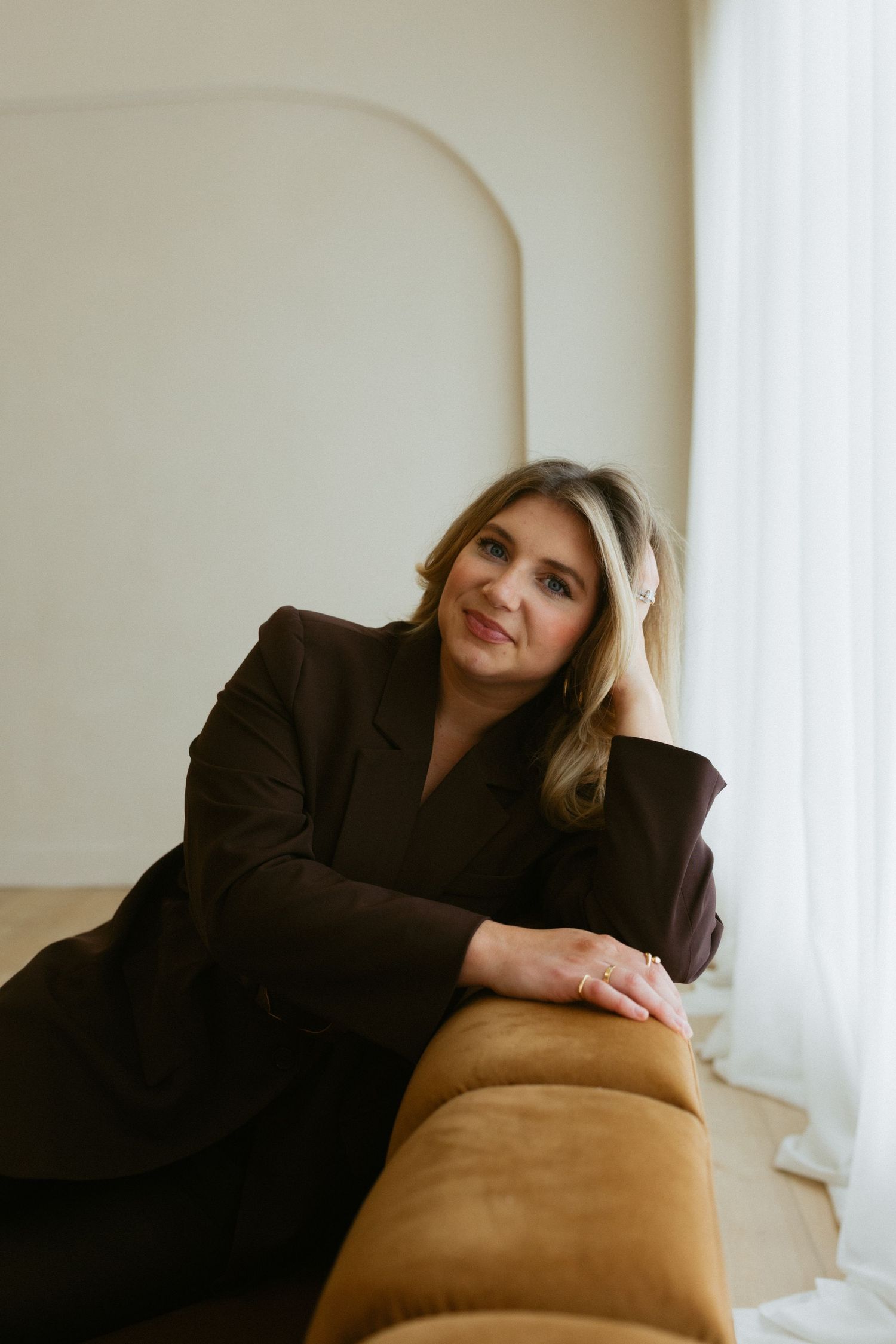 Person in tan hat and black sweater sits on white chair against gold curtain backdrop.