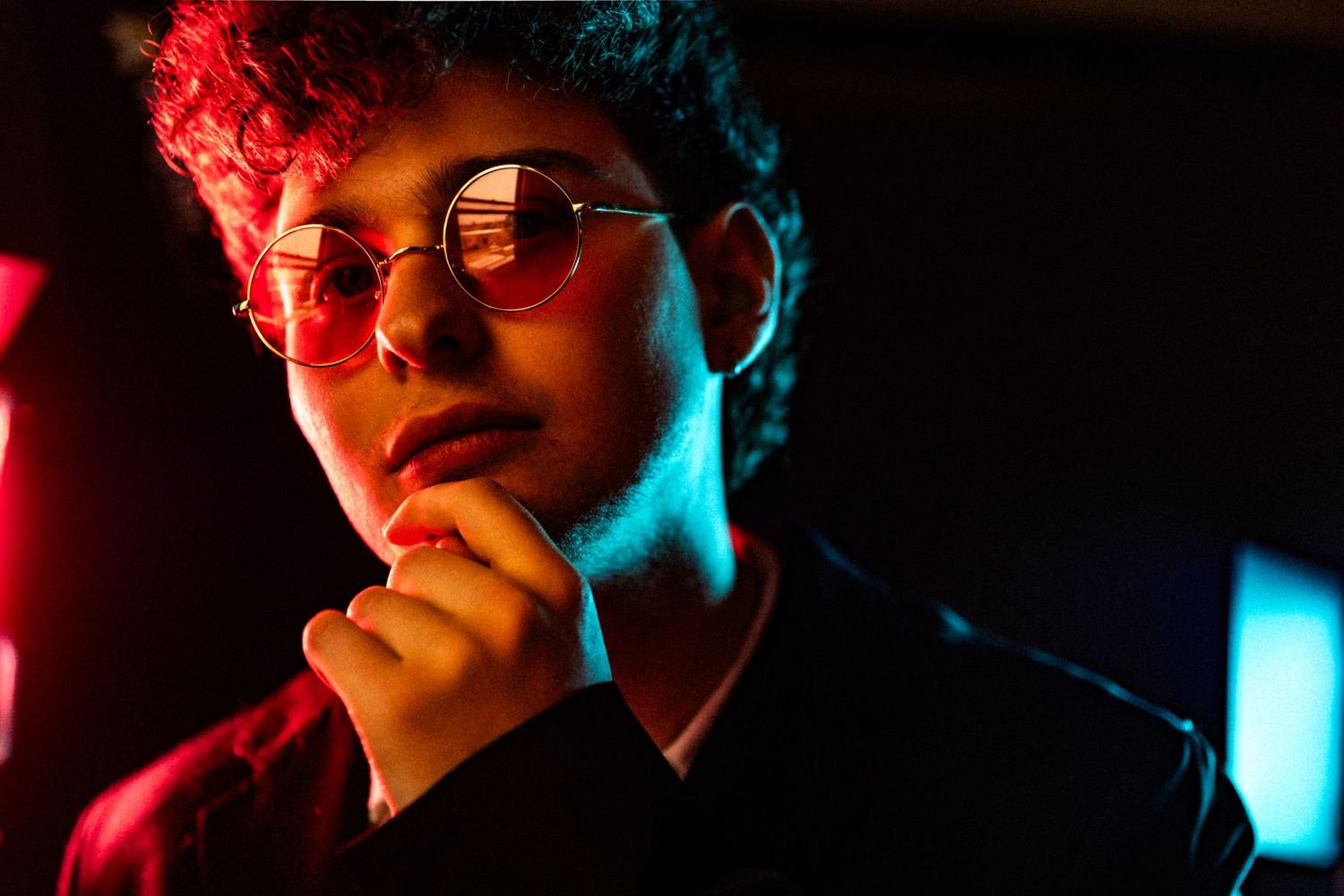 A moody portrait in neon lighting with red and blue tones showing a person wearing sunglasses in contemplative pose.