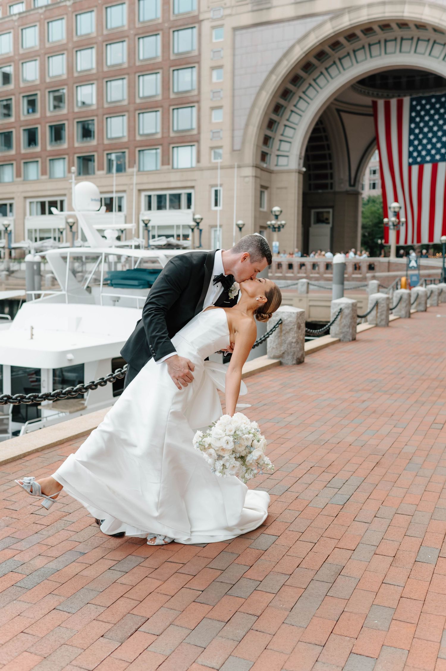 Romantic wedding couple performs traditional dip pose on red brick courtyard with American flag and boats in background.