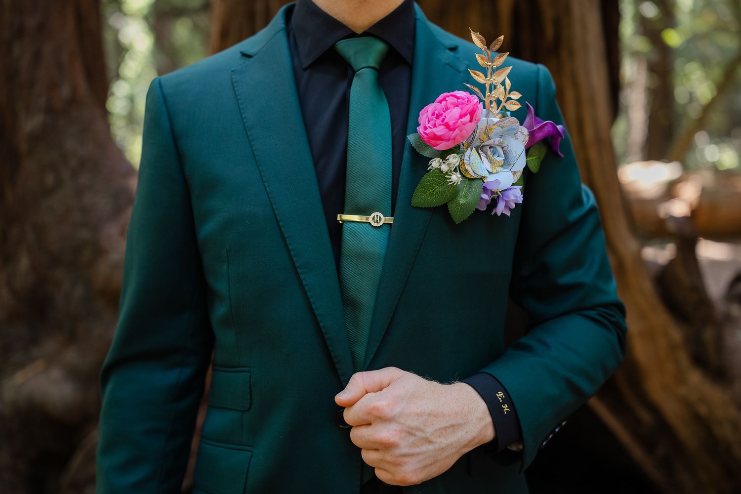 A person in a light gray suit poses outdoors on a path surrounded by trees and flowering bushes in a garden setting.