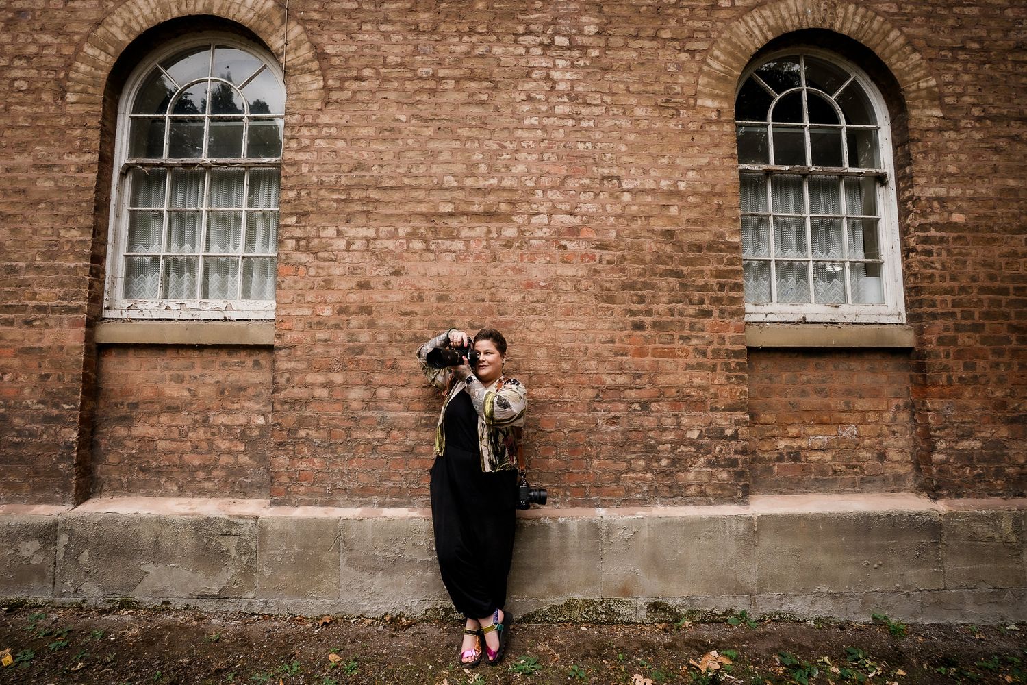 A figure in black stands against a rustic brick wall with arched windows in a vintage architectural setting.