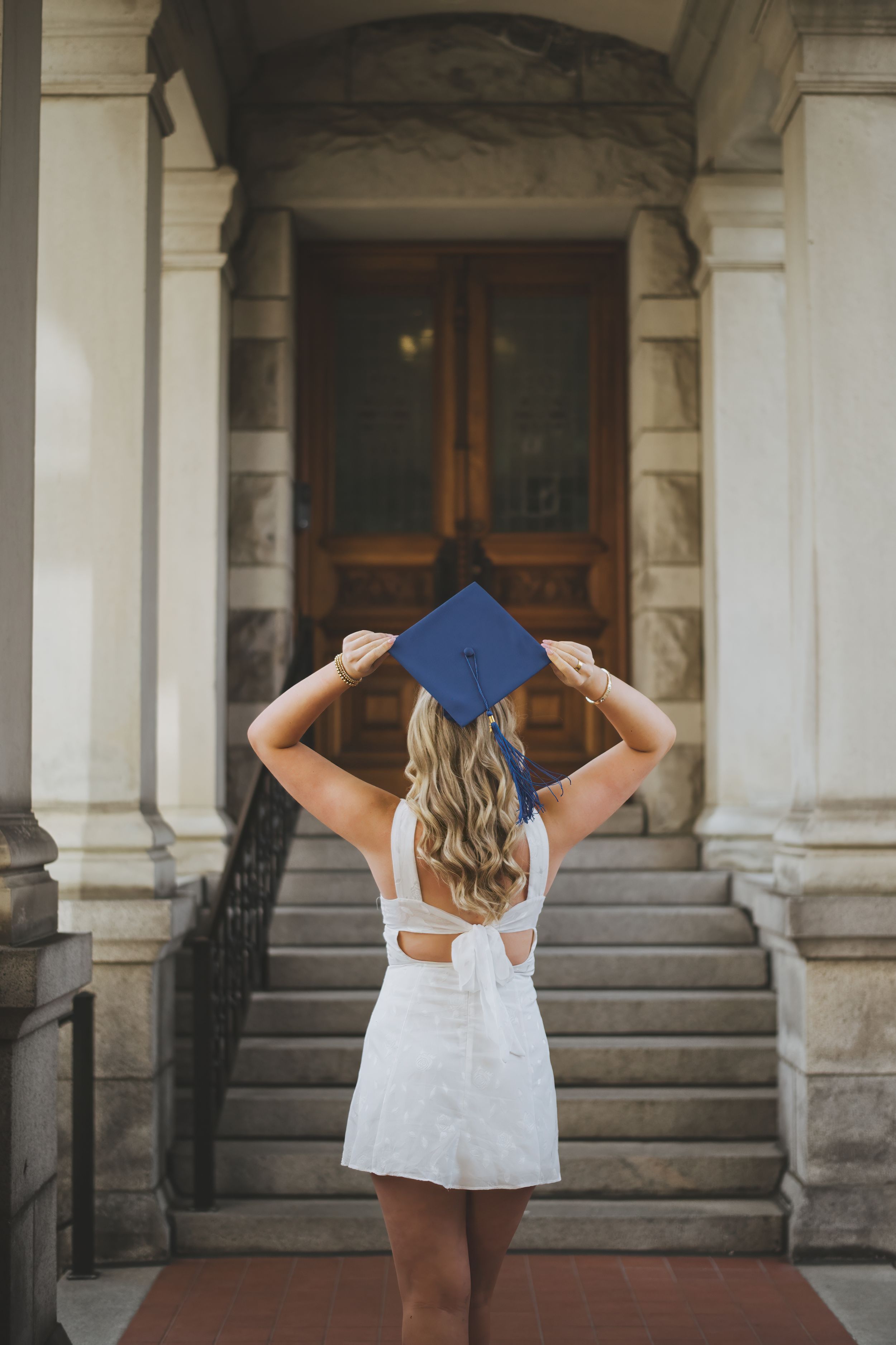 Graduate in white dress holding blue graduation cap stands in front of ornate building entrance.
