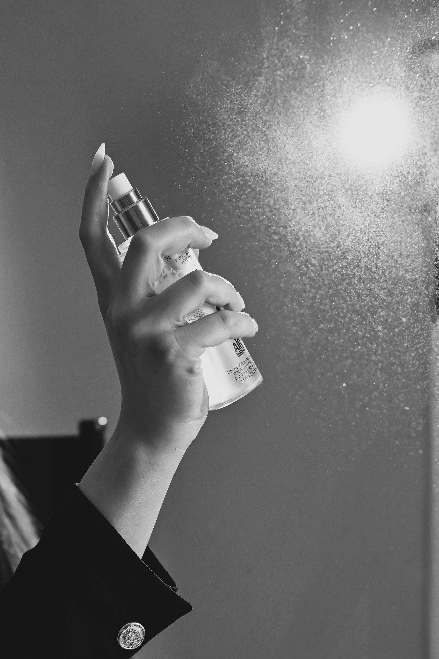 A hand sprays perfume in black and white lighting, creating a misty effect against a dark background.