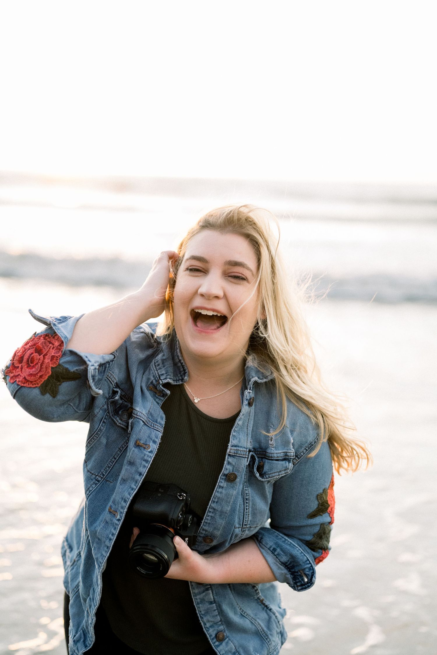 A person wearing a denim jacket and black top laughs joyfully while posing on a beach at sunset.