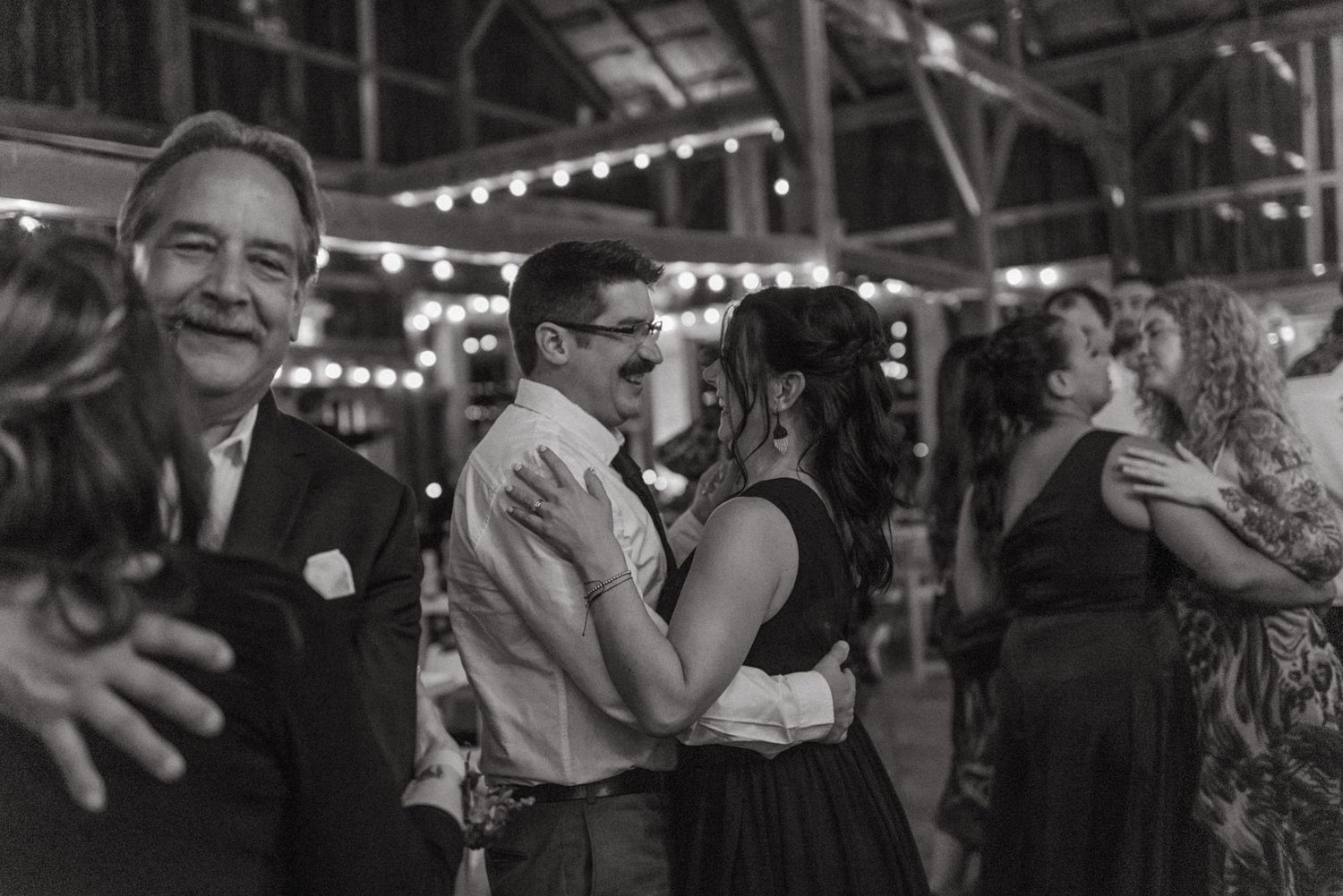 Black and white photo of couples dancing at an indoor event with string lights and rustic decor.