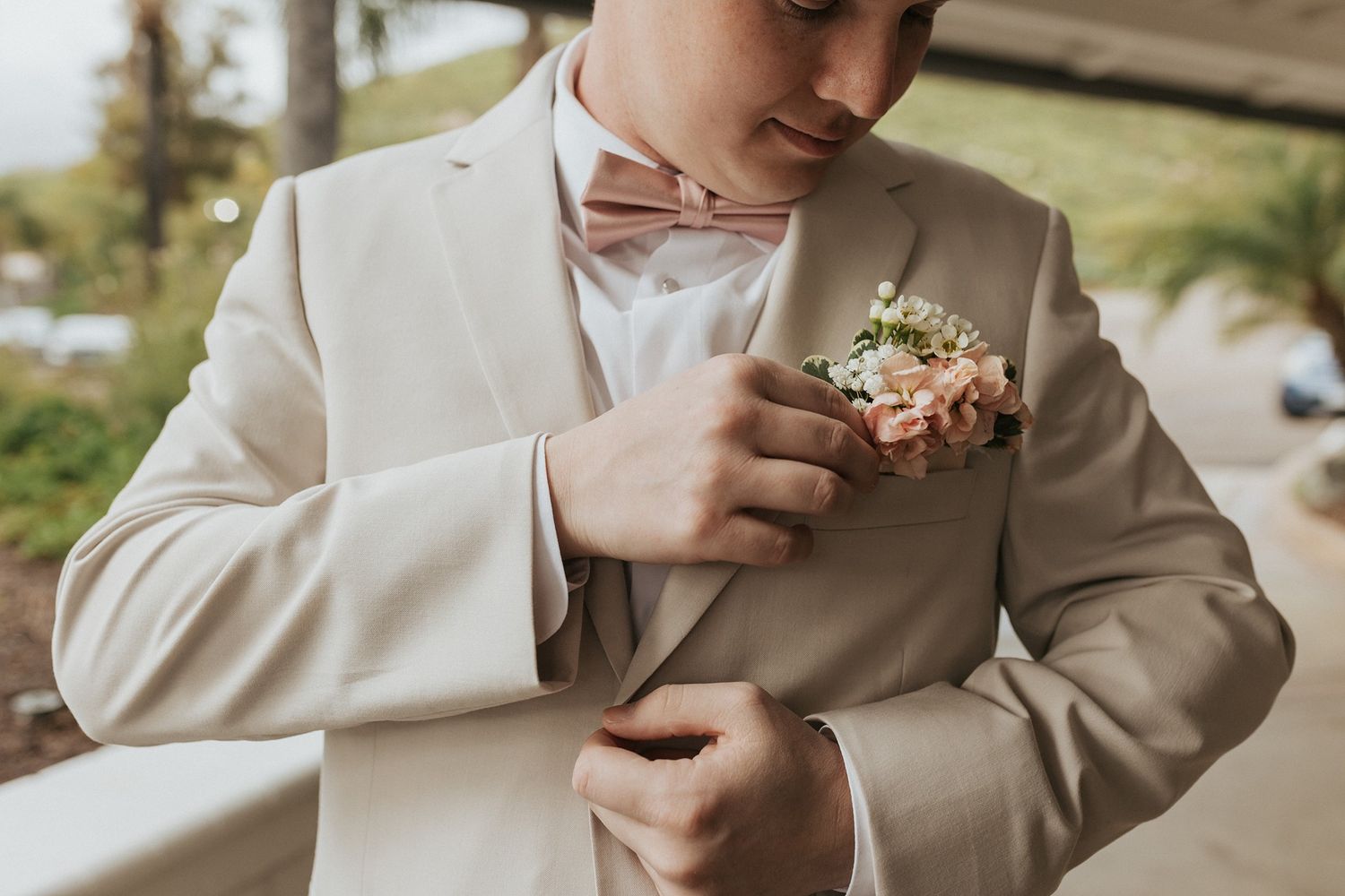 A groom in a beige suit adjusts his boutonniere with white flowers before the wedding ceremony.