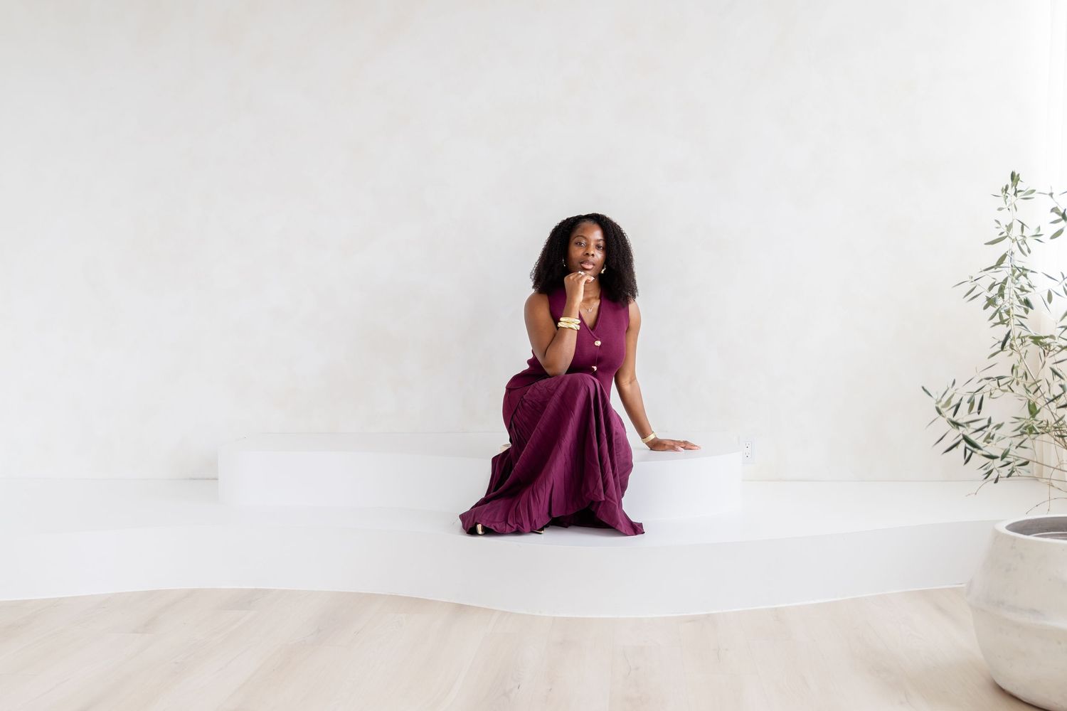 A person in a flowing burgundy dress poses gracefully against a white wall in a minimalist setting.